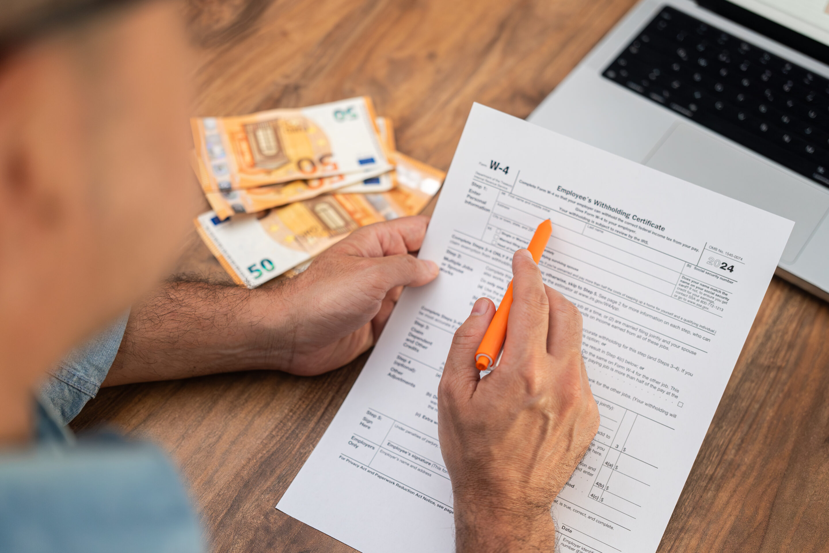 Man filling out W-4 tax form with euro banknotes and laptop on wooden desk Financial management and tax preparation process